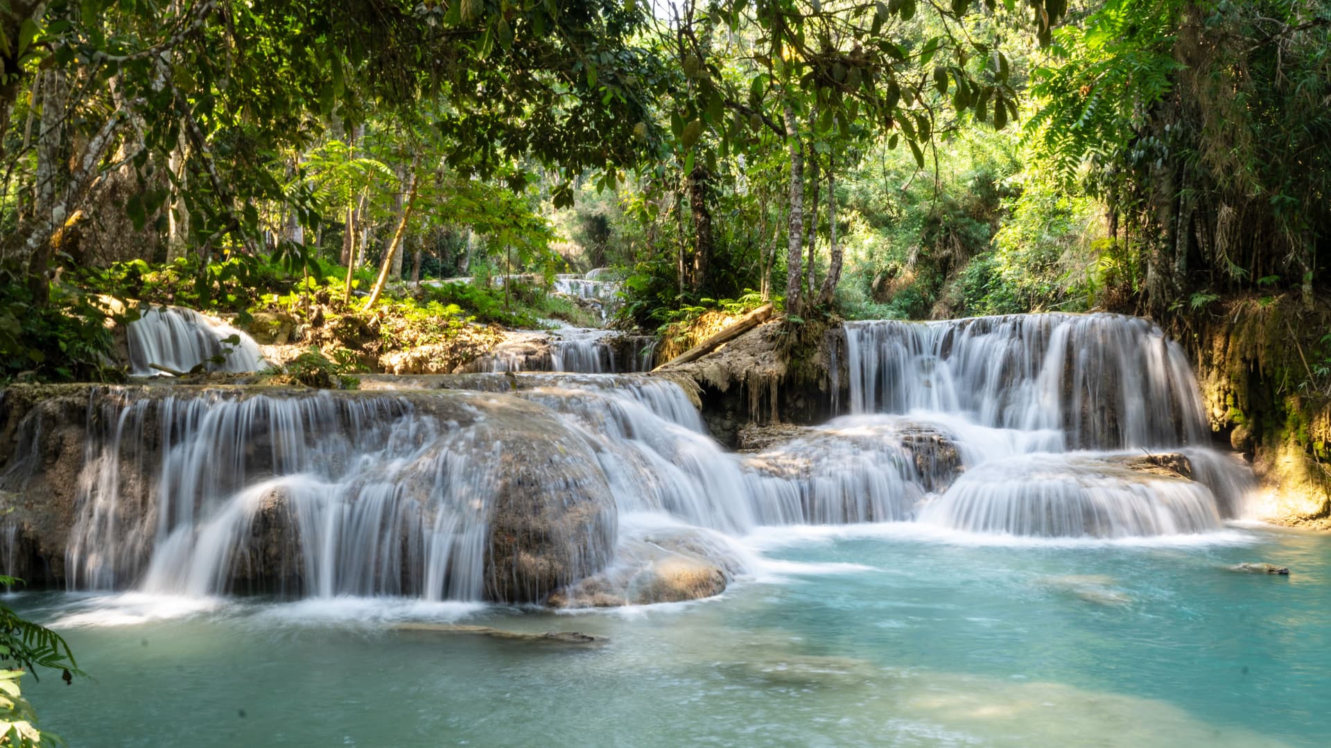 Tat Kuang Si Waterfalls in Laos, with multi tier cascades flowing over limestone into turquoise pools surrounded by forest.