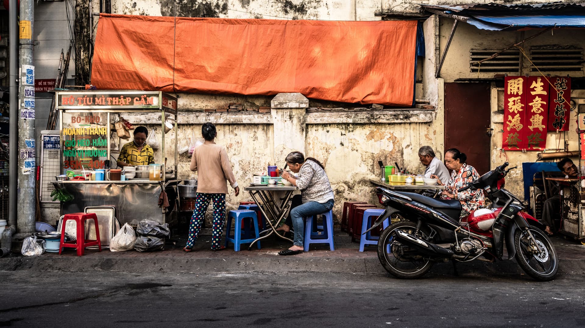 Street food stall in Vietnam with locals eating on small plastic stools and tables beside a worn wall, with a motorbike parked in front.