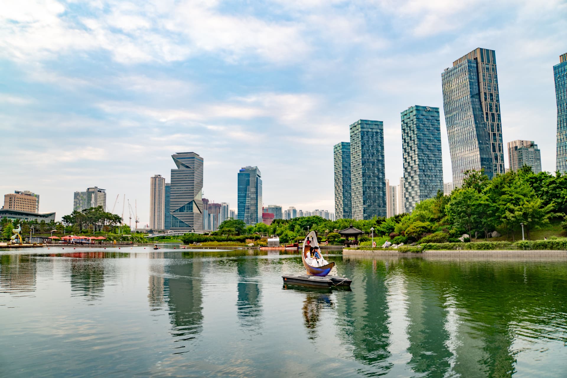 Modern South Korea city skyline reflected on a calm lake with a small boat in the foreground and a green park along the waterfront.