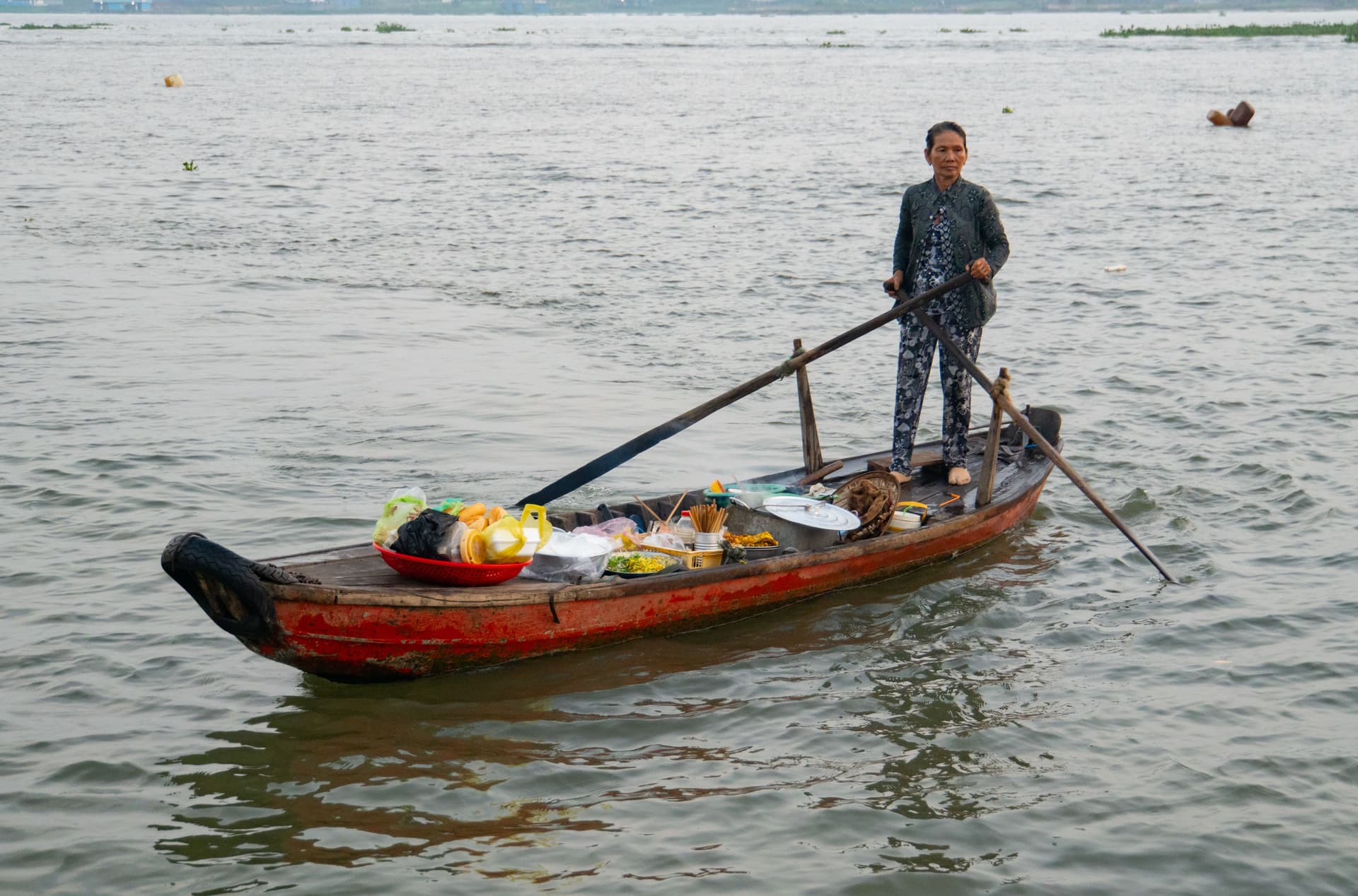 Woman standing on a small wooden boat in Vietnam, rowing across a river with baskets of food and cooking pots onboard.