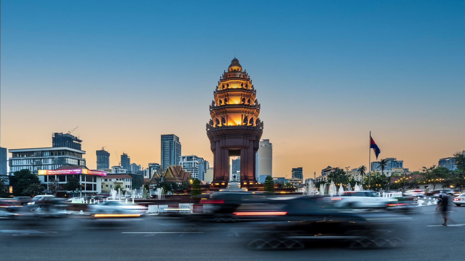 Independence Monument in Phnom Penh, Cambodia glowing at dusk with city buildings in the background and blurred traffic circling the roundabout.
