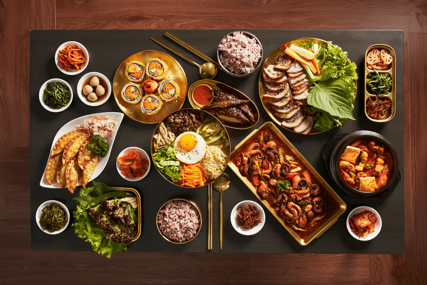 Overhead view of a Korean meal spread with banchan side dishes, kimchi, rice, kimbap, bibimbap, sliced pork, dumplings, and spicy tofu stew on a table.